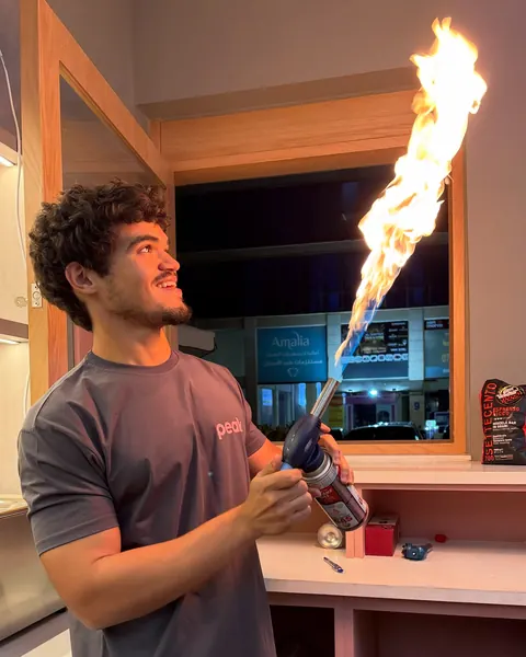 Hasan, the owner, in a grey peak tee behind the counter, holding up a lit kitchen torch.